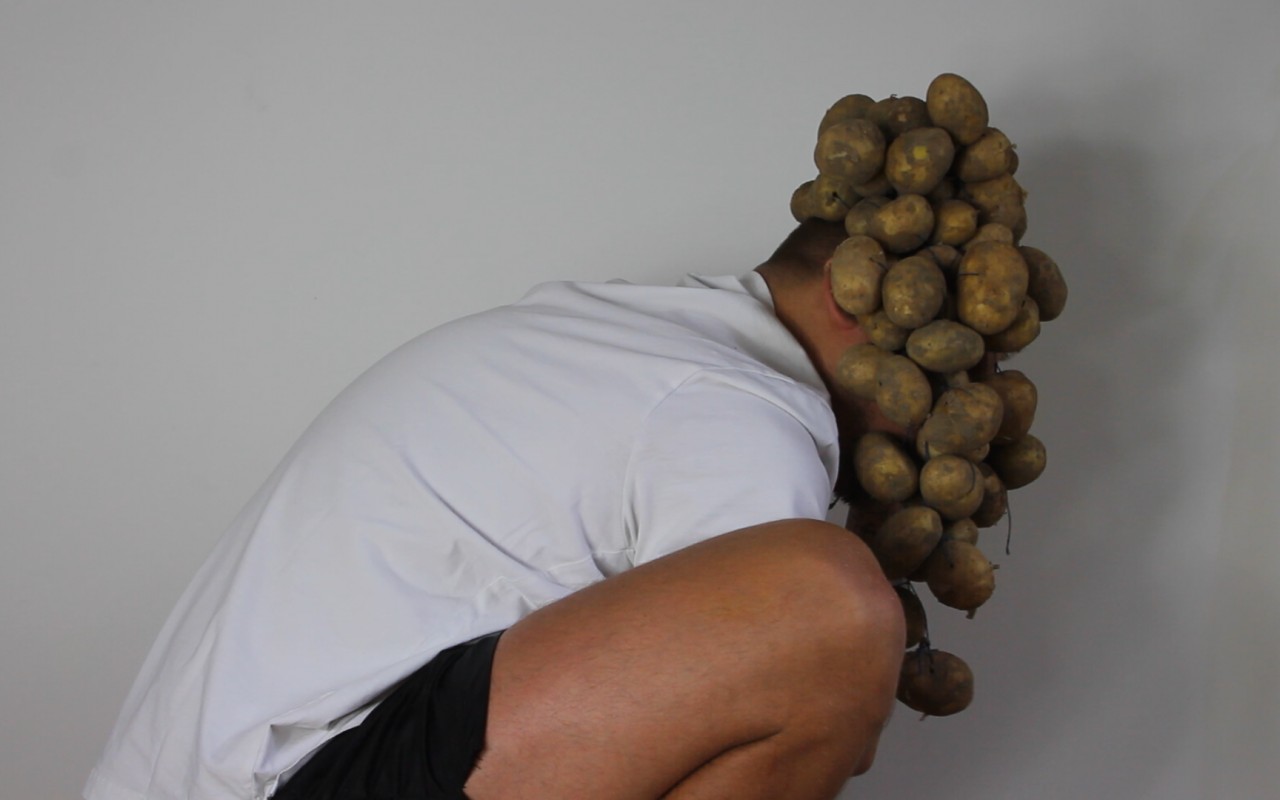 mężczyzna w przykucu, pokazany bokiem, na głowie ma ziemniaki