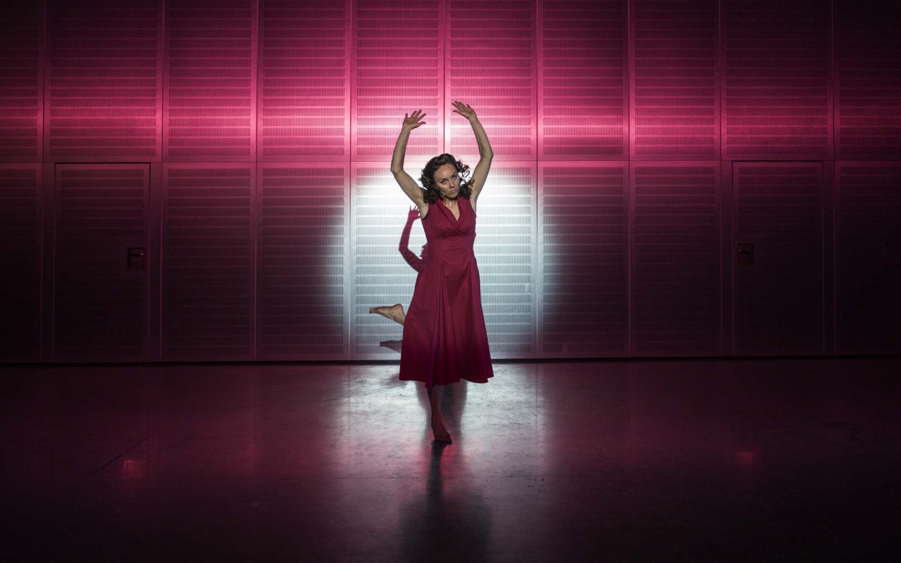 Kobieta ubrana w bordową sukienkę stoi w pozie baletowej z uniesionymi rękami. Na nią skierowane jest światło sceniczne.