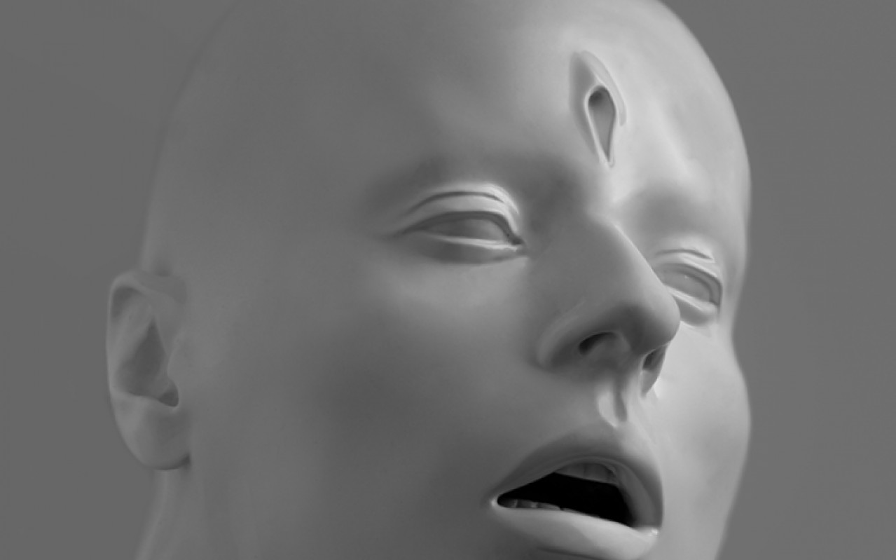 Rzeźba głowy wraz z dłońmi. Łysa głowa oparta brodą o dłonie z otwartymi ustami, na czole podłużny otwór.