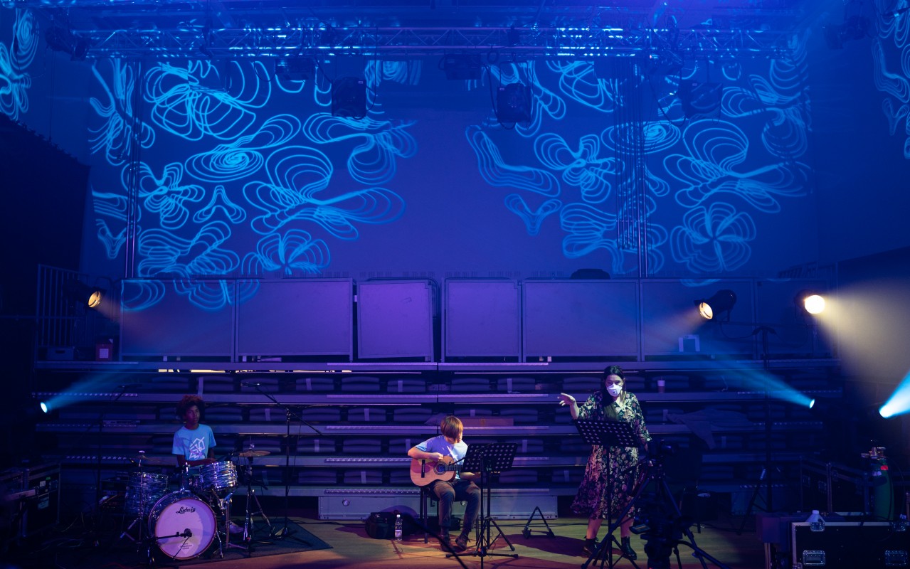 trzy osoby grające na instrumentach w sali oświetlonej zdobnie na niebiesko