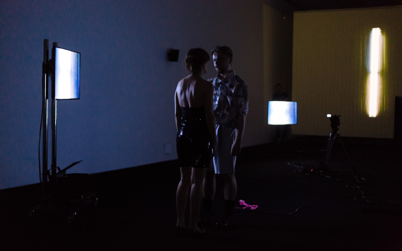 kobieta i mężczyzna stoją na przeciw siebie w ciemnym pokoju, oświetla ich blask z ekranów