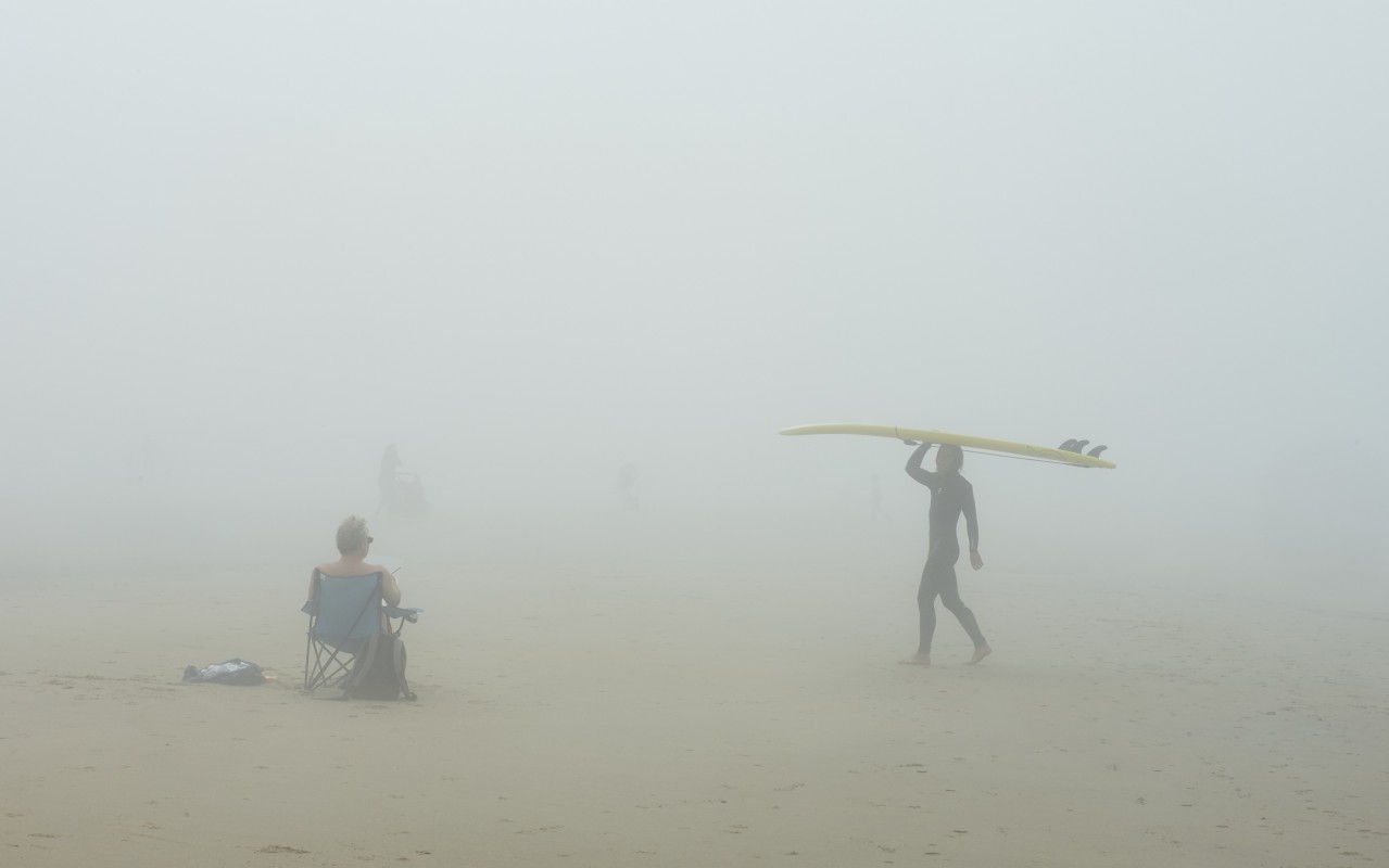Na zamglonej plaży siedzi człowiek na krześle tyłem do patrzącego, od strony morza idzie surfer z deską nad głową