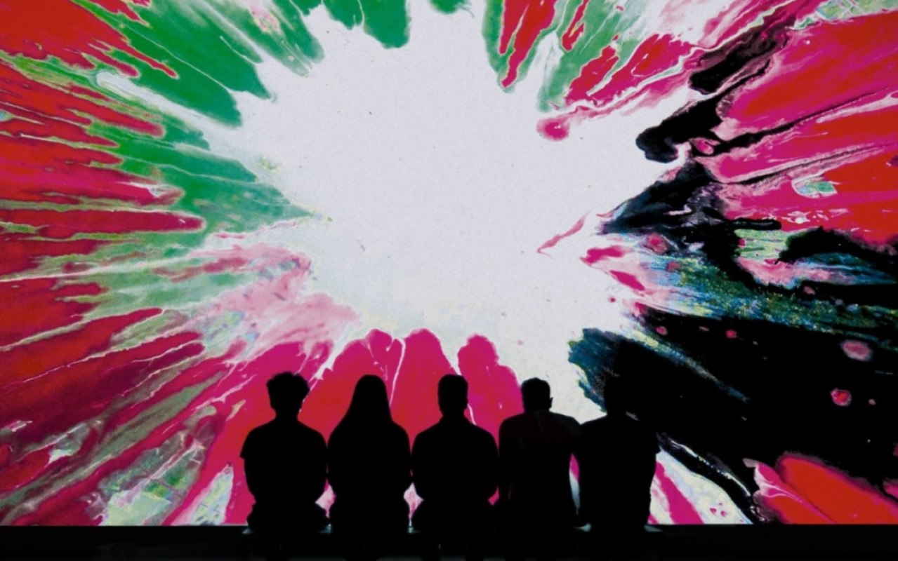 Pięć osób siedzi tyłem, widać tylko ich ciemne sylwetki na tle ekranu z kolorowym rozbryzgiem