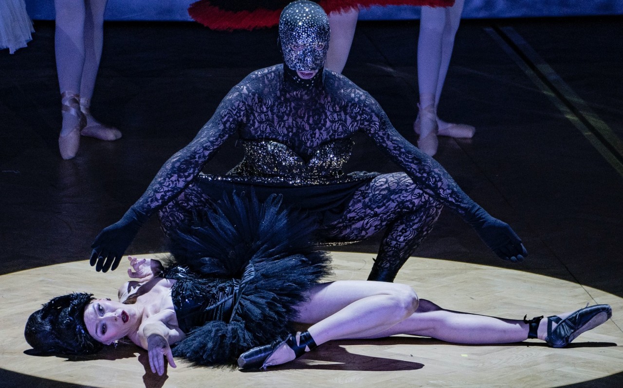 W świetle reflektora na podłodze leży kobieta w czarnym tutu baletnicy, nad nią przykucnięty jest człowiek w masce i koronkowym kombinezonie