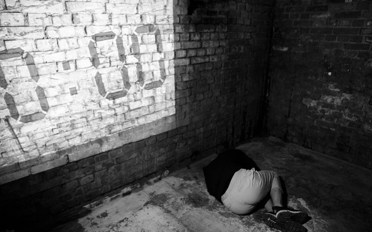Czarno-białe zdjęcie z postacią lezącą na ziemi w pustym, surowym pomieszczeniu. Na ścianie wyświetla się godzina zegarowa 00:00.