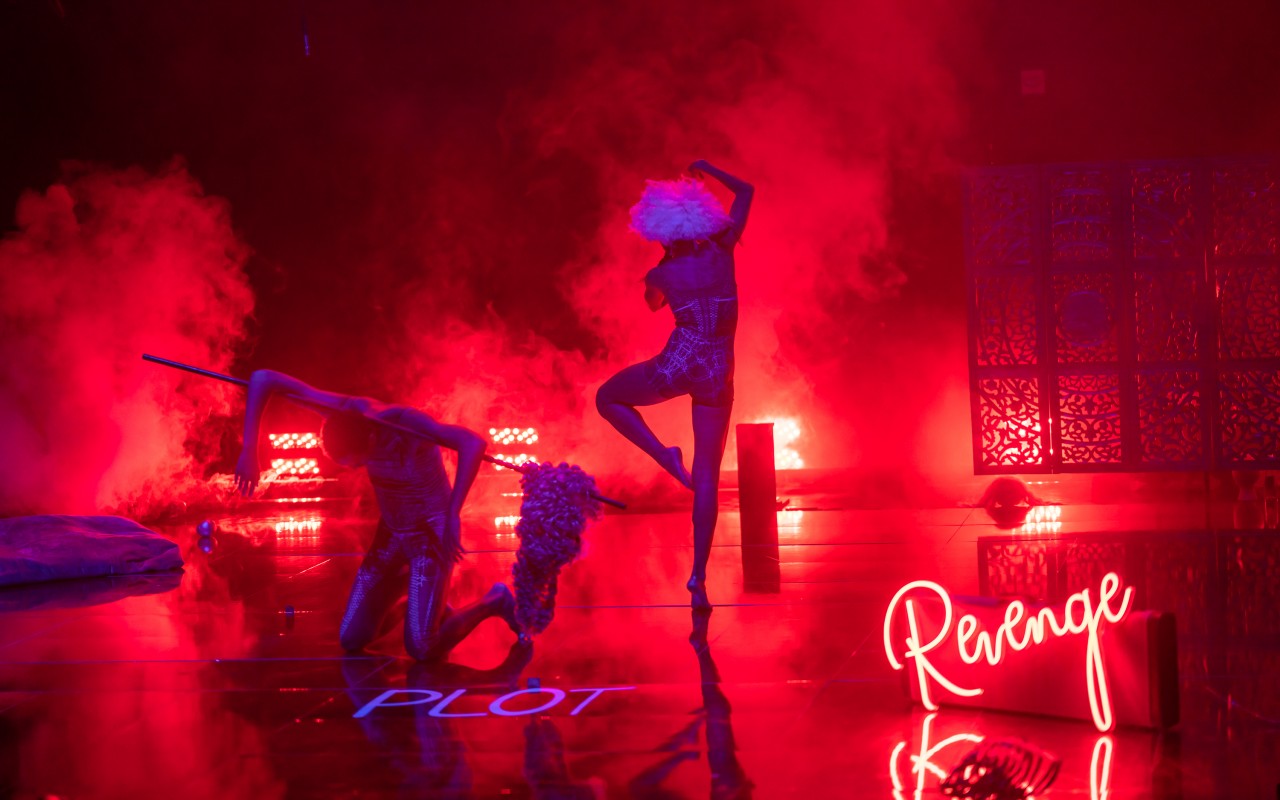 Na scenie oświetlonej czerwono-niebieskim światłem widać dwie postaci w ruchu, na podłodze leżą pojedyncze przedmioty m.in. peruka