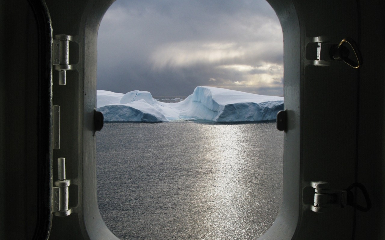 Fotografia przedstawia otwarte okno kajuty, widok z wewnątrz, przez okno widać ocean i lodowy pokryty śniegiem masyw, na oknie leżą naszyjnik i pierścionek z brylantem