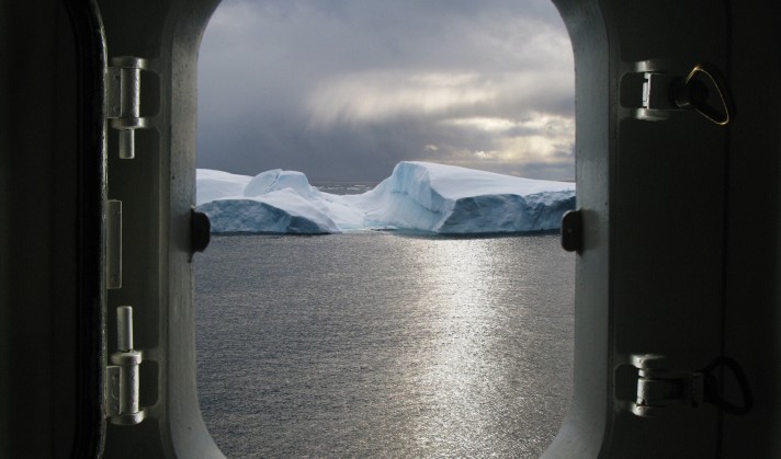 Fotografia przedstawia otwarte okno kajuty, widok z wewnątrz, przez okno widać ocean i lodowy pokryty śniegiem masyw, na oknie leżą naszyjnik i pierścionek z brylantem