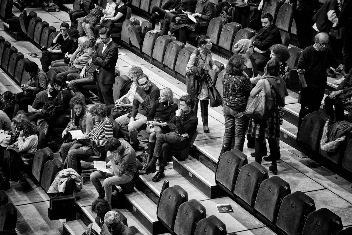 Fotografia czarno-biała przedstawiająca widownię teatralną, niektórzy widzowie siedzą czekając na spektakl, inni stoją pomiędzy rzędami i rozmawiają. Po środku wśród innych siedzi na fotelu teatralnym Krzysztof Warlikowski
